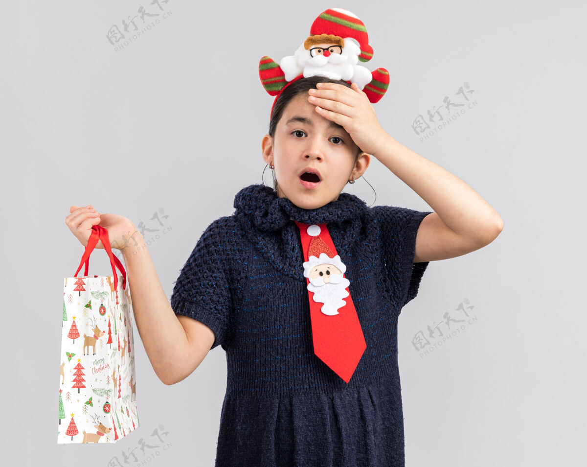 包穿着针织连衣裙的小女孩头上系着一条红色领带 头上戴着一个有趣的圣诞边 手里拿着一个装有圣诞礼物的纸袋 看上去既惊讶又惊奇头惊讶惊讶