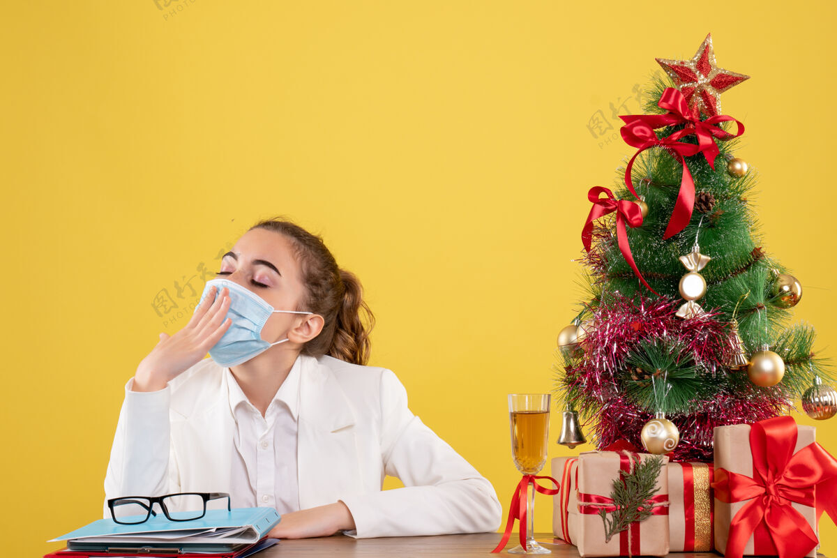 商务正面图女医生戴着防护面具坐在黄色背景上打着哈欠 手里拿着圣诞树和礼盒肖像打哈欠圣诞节