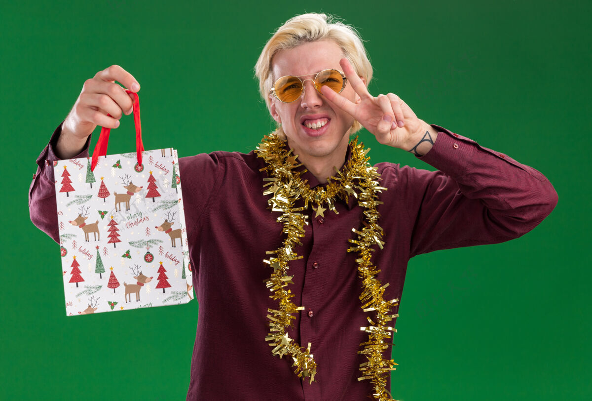 圣诞节快乐的年轻金发男子戴着眼镜 脖子上戴着金箔花环 手里拿着圣诞礼品袋 在绿色的墙上做着和平的手势周围男人和平