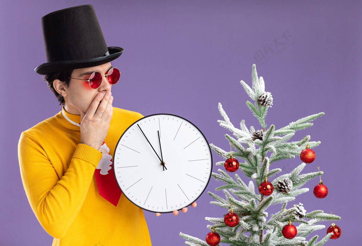 圣诞节一个穿着黄色高领毛衣 戴着黑帽子的年轻人拿着挂钟 站在紫色墙上的圣诞树旁 好奇地看着它男人时钟帽子