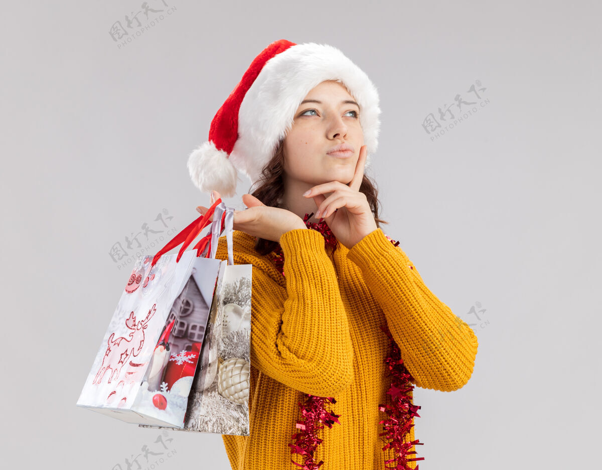 圣诞快乐体贴的年轻斯拉夫女孩戴着圣诞帽 脖子上戴着花环 手放在下巴上 手里拿着纸礼包 看着旁边圣诞老人纸周到