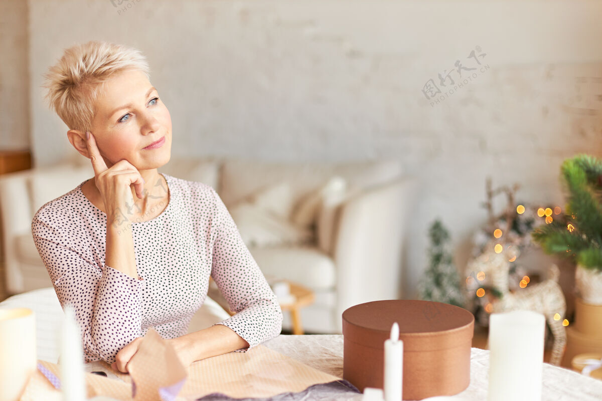 女人一位体贴的中年女性的画像 穿着优雅的衣服 坐在装饰着花环的房间里的桌子旁 神情沉思 把食指放在头上 想着如何为家人包装礼物包装惊喜手工制作
