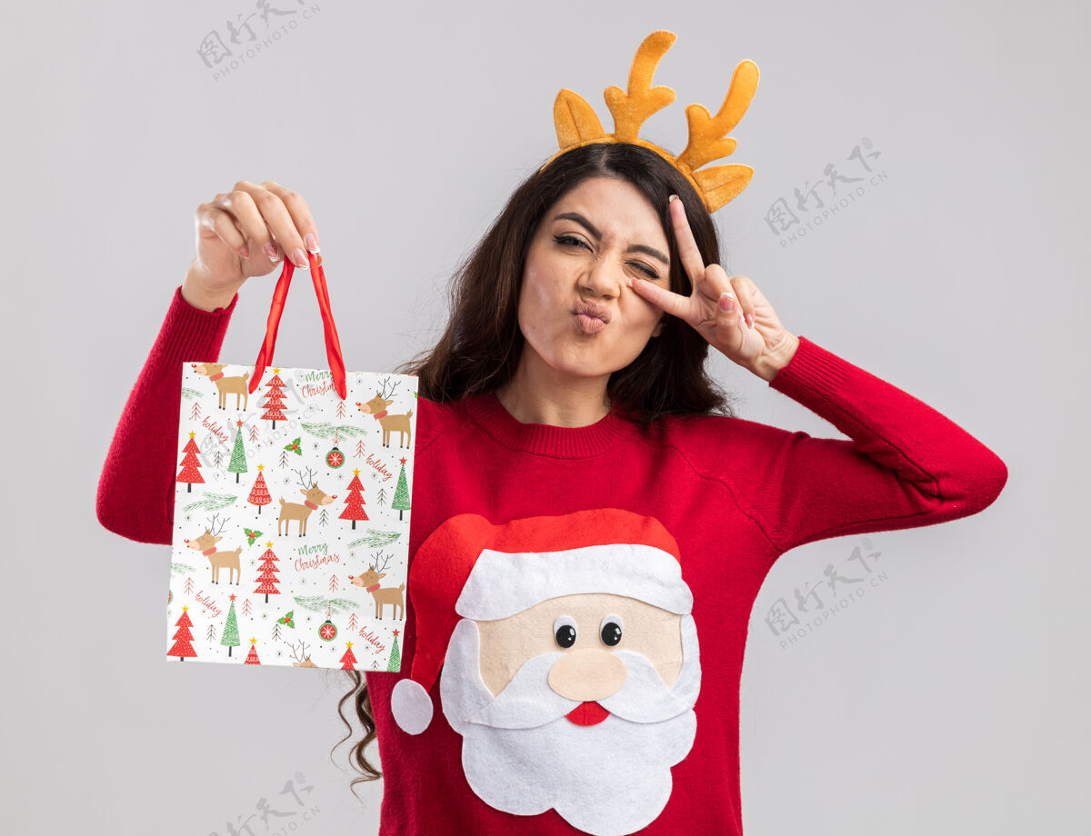 眨眼自信的年轻漂亮女孩戴着驯鹿鹿角头带和圣诞老人毛衣拿着圣诞礼品袋看着展示着v字符号近眨眼自信年轻圣诞快乐