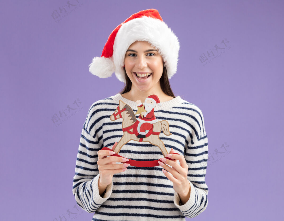马快乐的年轻白人女孩戴着圣诞帽拿着圣诞老人在摇马上装饰新年紫色快乐