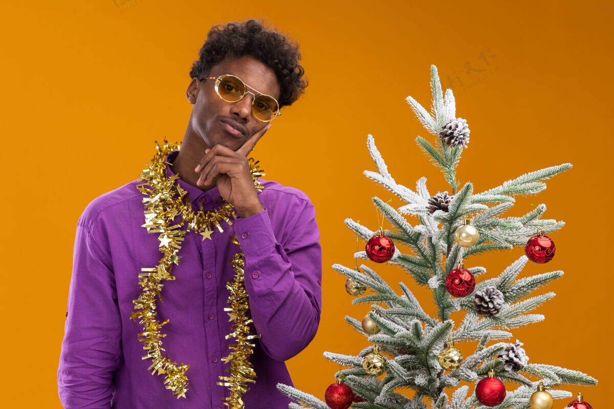 站体贴的美国黑人年轻人戴着一副眼镜 脖子上戴着金箔花环 站在橙色背景的装饰过的圣诞树旁装饰金属片近
