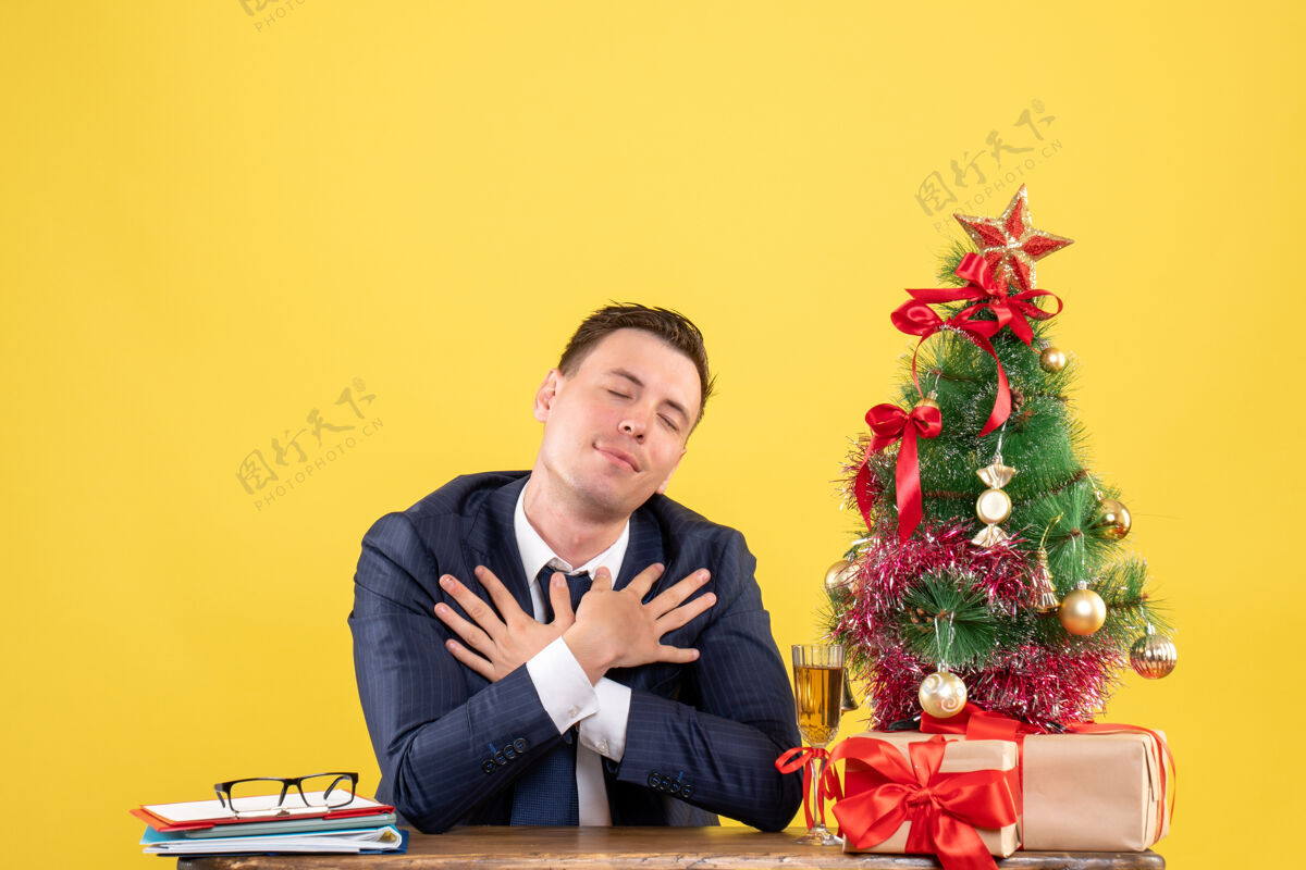 商人前视图满意的人把他的手放在胸前 坐在圣诞树附近的桌子和黄色的礼物放西装圣诞节