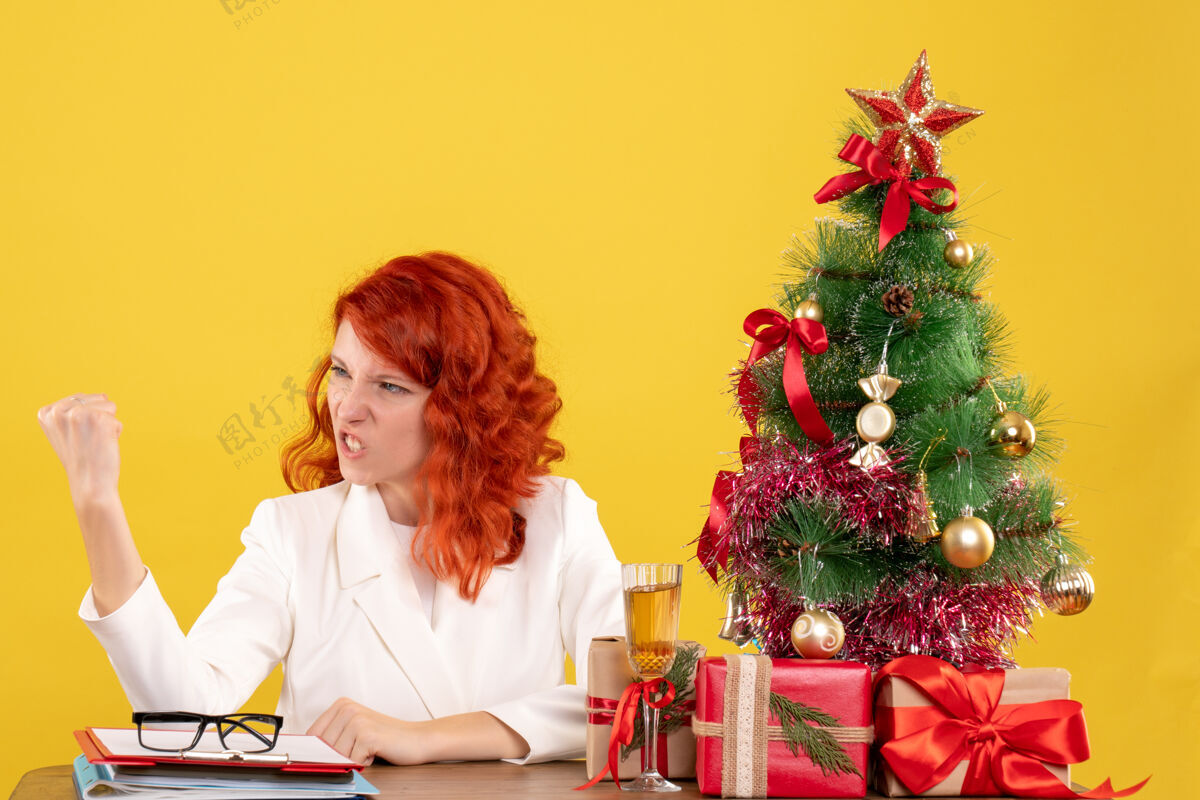 微笑正面图：女医生坐在桌子后面 黄色背景上有圣诞礼物桌子威胁女性