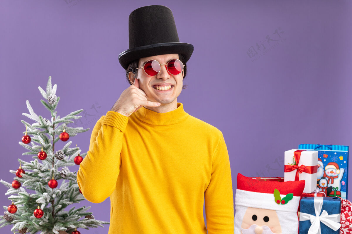 做一个穿着黄色高领毛衣 戴着黑色帽子 戴着眼镜 面带微笑的年轻人站在圣诞树旁做着“打电话给我”的手势 在紫色的背景下送礼物树相机帽子