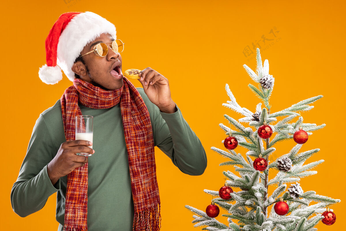 饼干一个戴着圣诞帽 围着围巾 拿着饼干 拿着牛奶的非洲裔美国人站在橙色背景下的圣诞树旁美国人非洲围巾