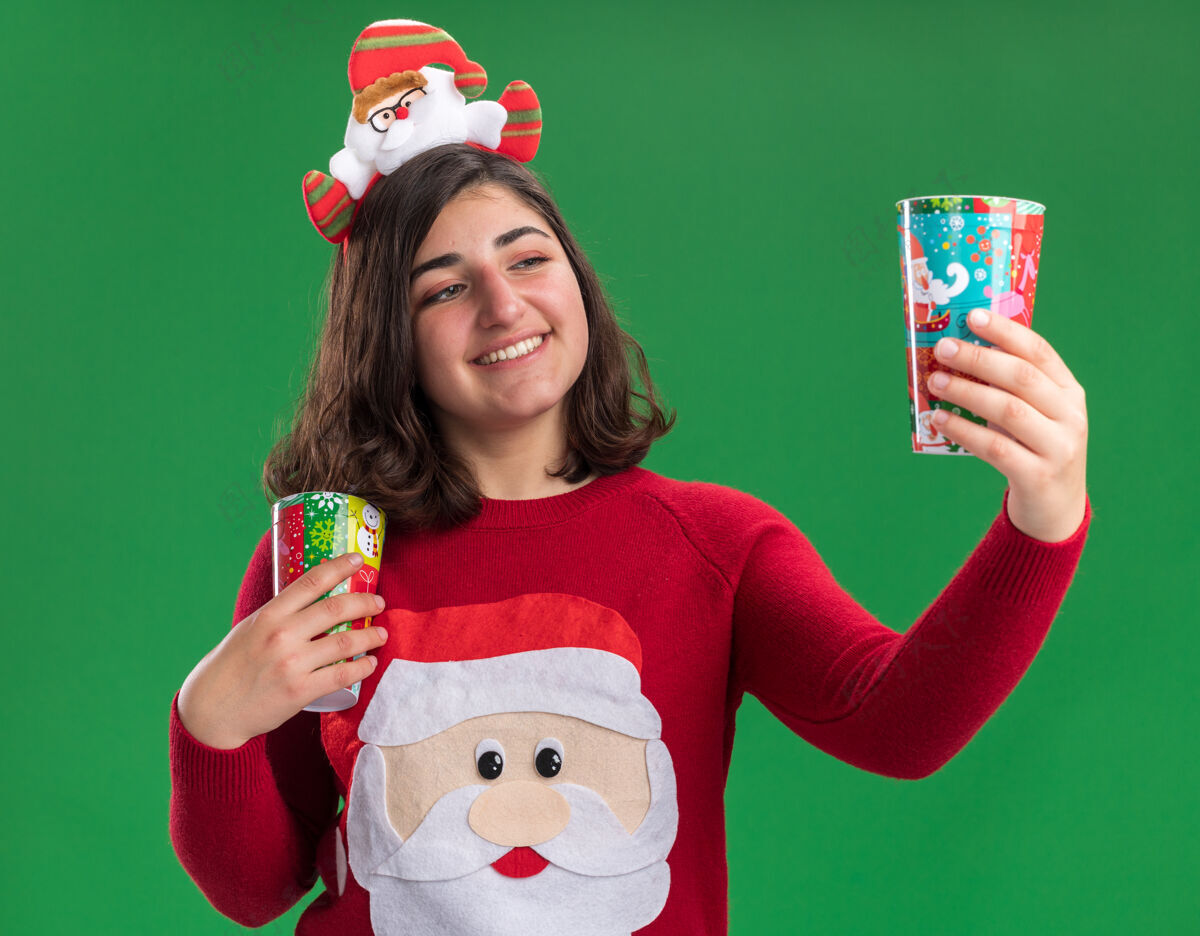 圣诞节快乐的小女孩穿着圣诞毛衣 戴着圣诞帽 手里拿着五颜六色的纸杯 面带微笑地看着它站在绿色的墙上穿着站着五颜六色