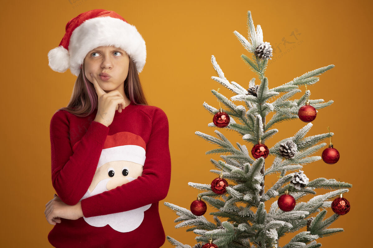 思想小女孩穿着圣诞毛衣 戴着圣诞帽 在橙色背景下的圣诞树旁 神情沉思地抬头望去小毛衣站着