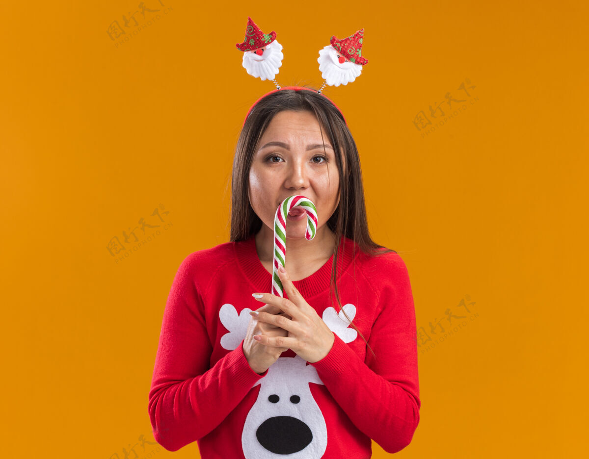 毛衣年轻的亚洲女孩戴着圣诞发箍和毛衣拿着圣诞糖果隔离在橙色的墙上女人年轻糖果