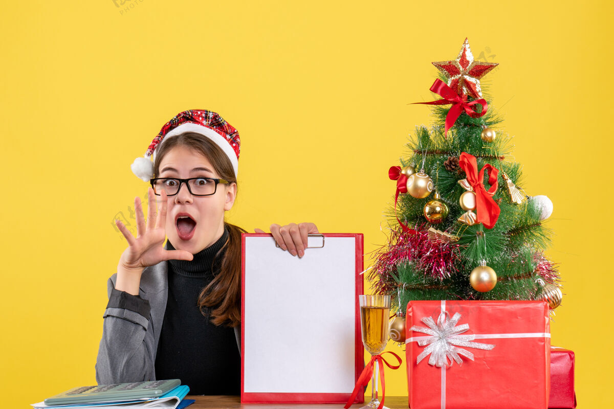女孩前视图好奇的女孩戴着圣诞帽坐在桌旁张嘴圣诞树和礼物鸡尾酒庆典喜剧演员好奇的女孩