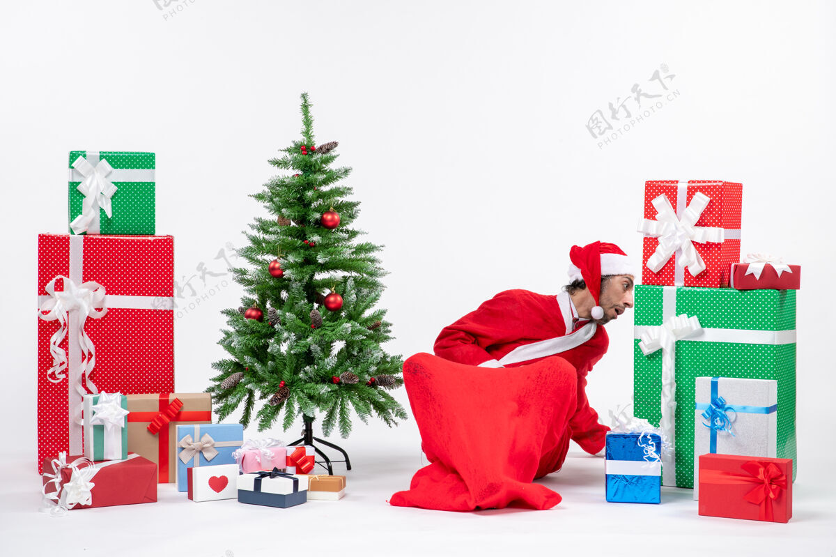 地面年轻人打扮成圣诞老人 拿着礼物和装饰好的圣诞树坐在地上找东西礼物看东西