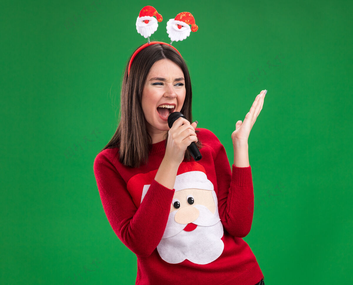 穿年轻漂亮的白人女孩穿着圣诞老人的毛衣和头带拿着麦克风看着一边保持手在空中唱歌隔离在绿色的墙壁上复制空间圣诞老人空气保持