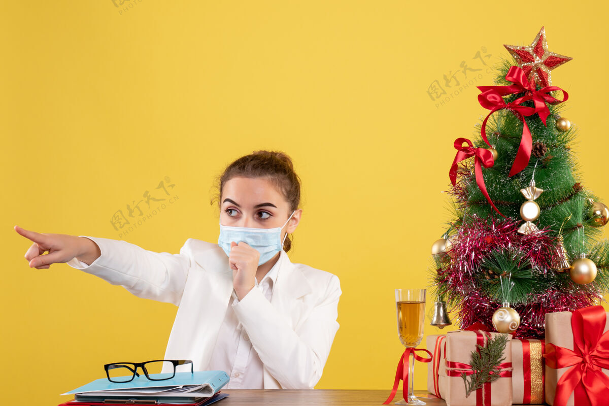男性正面图：戴着防护面具坐在黄色桌子上的女医生带着圣诞树和礼品盒外套防护实验室