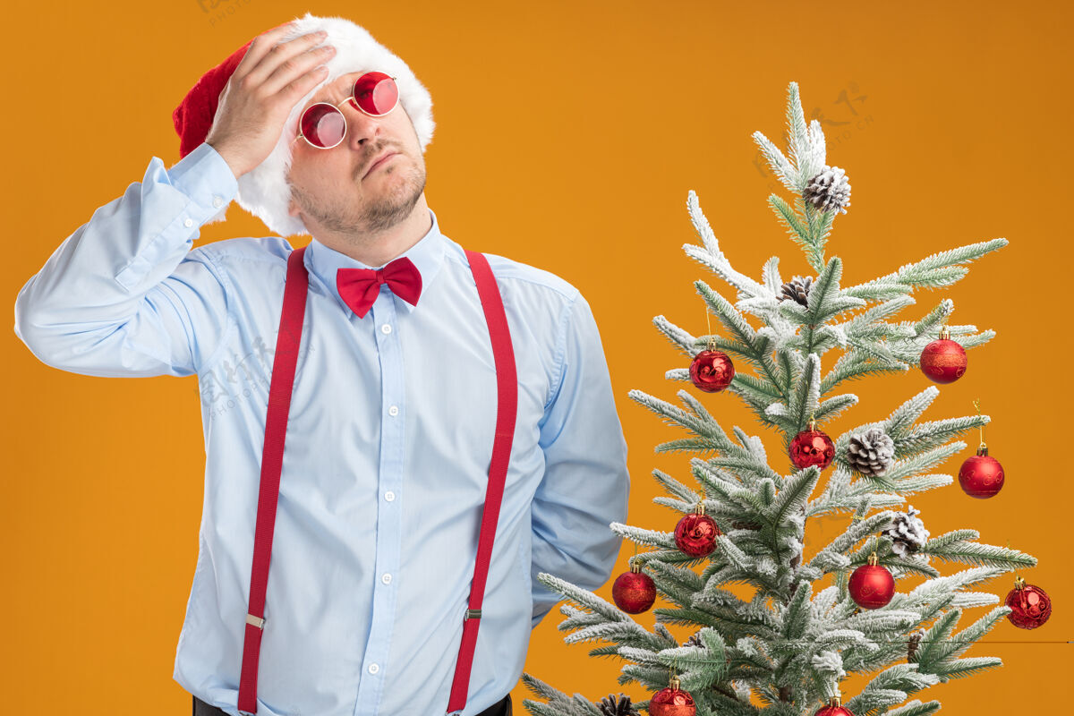 男人一个戴着圣诞帽 戴着红眼镜 系着吊带领结的年轻人站在圣诞树旁 用手捂着头望着橘色的墙上 困惑地抬起头来圣诞老人帽子穿着
