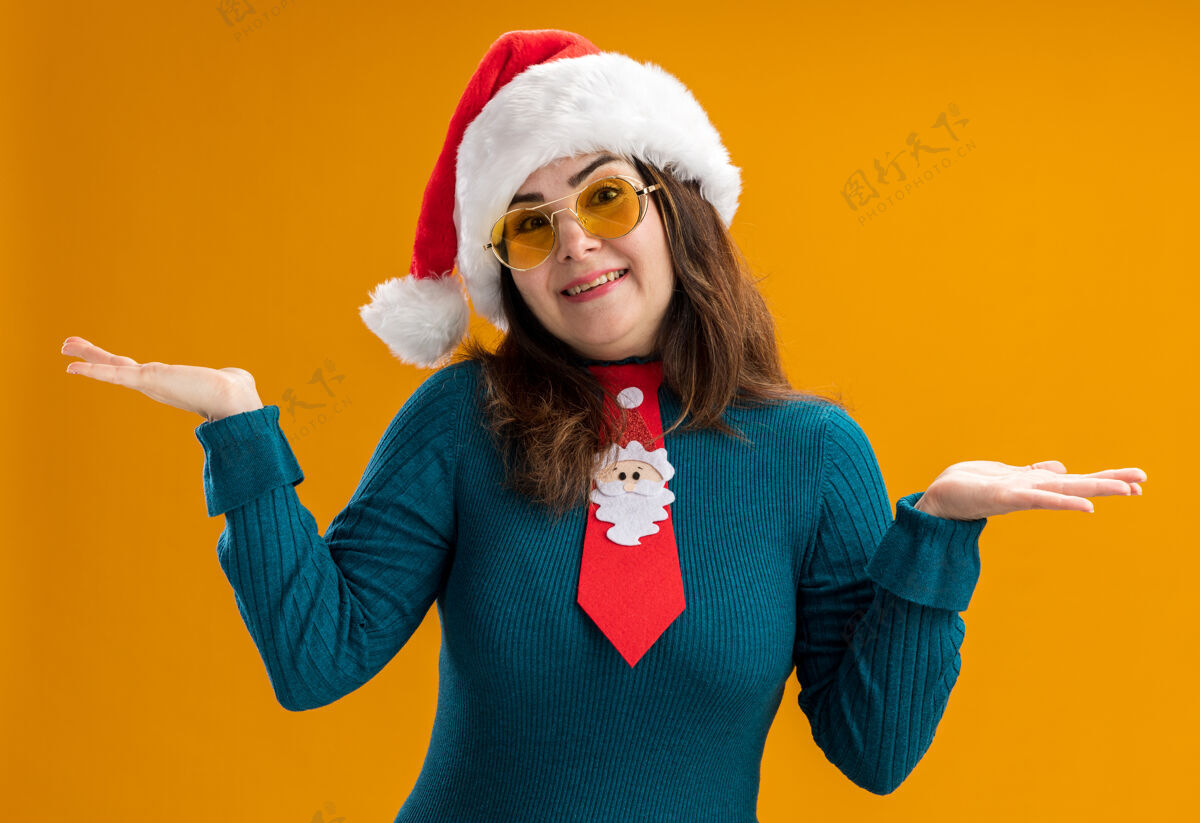 眼镜戴着太阳眼镜 戴着圣诞帽 打着圣诞领带 双手张开的微笑的成年白人妇女拿着橙色成人