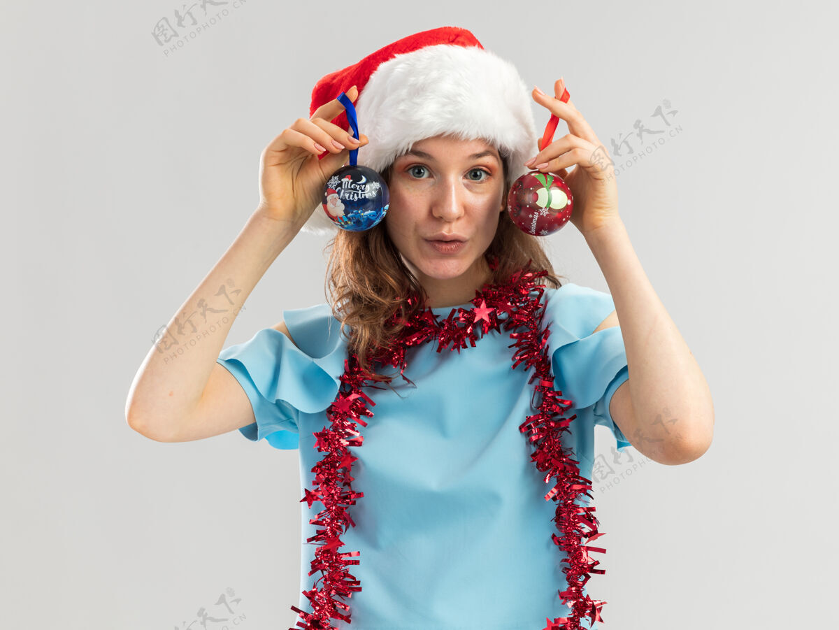 顶身着蓝色上衣 头戴圣诞老人帽 脖子上戴着金属丝的快乐年轻女子 手里拿着圣诞球 自信地微笑着自信圣诞快乐女人
