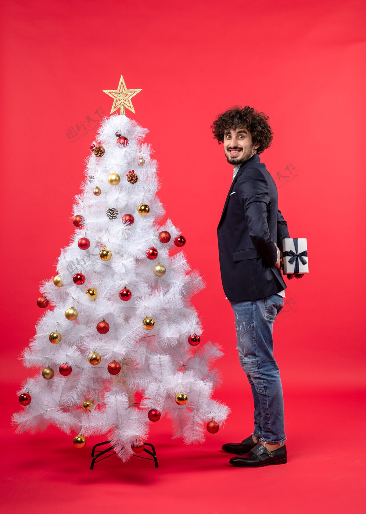 庆祝快乐的年轻人拿着他的礼物站在红色的右侧装饰的白色圣诞树旁年轻人礼物圣诞节
