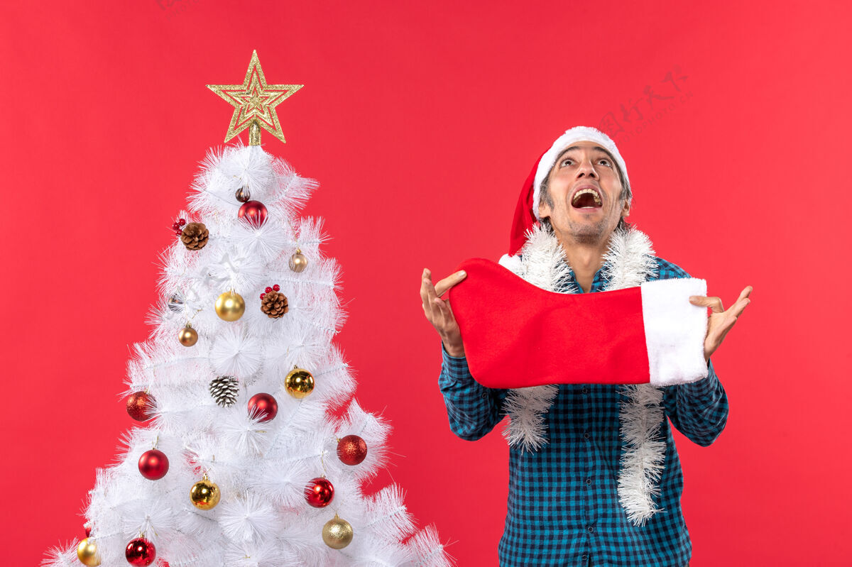 圣诞情绪激动的年轻人戴着圣诞老人的帽子 穿着蓝色的衬衫 手里拿着圣诞袜子 往上看帽子圣诞老人庆祝