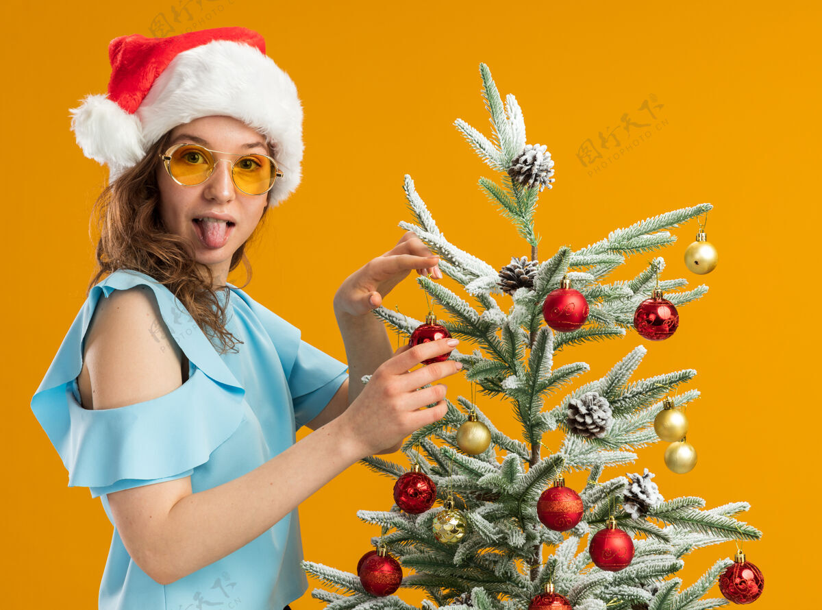 年轻穿着蓝色上衣 戴着圣诞帽 戴着黄色眼镜装饰圣诞树的快乐而积极的年轻女子伸出舌头看着站在橙色背景上的摄像机帽子站立装饰