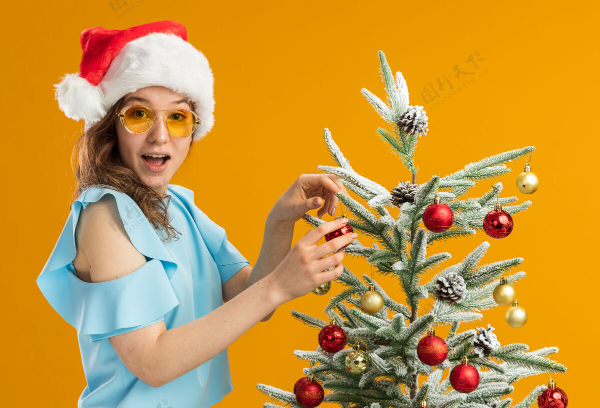 惊喜一个穿着蓝色上衣 戴着圣诞帽 戴着黄色眼镜 站在橙色背景下装饰圣诞树的快乐而惊讶的年轻女子帽子站着快乐