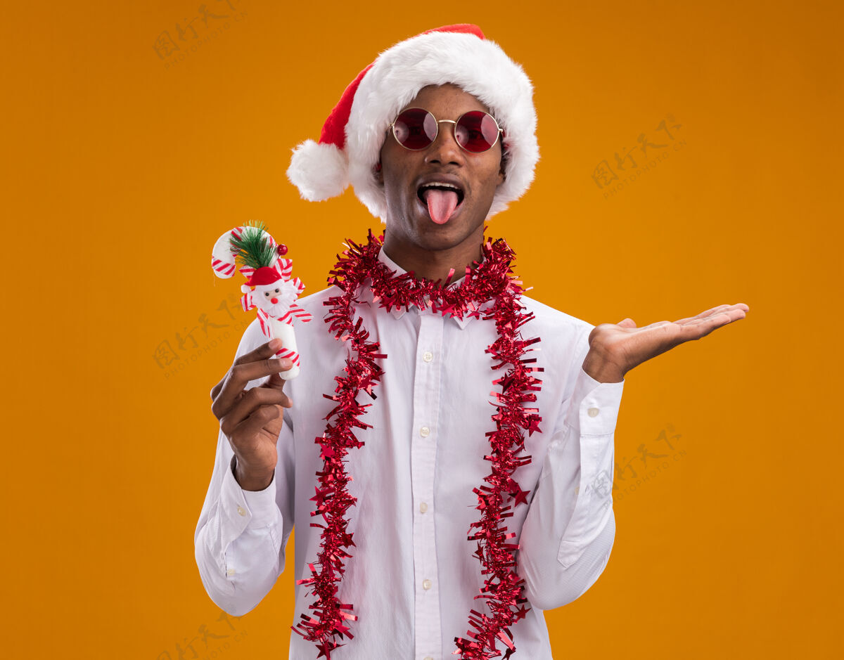 年轻顽皮的美国黑人年轻人 戴着圣诞帽 戴着眼镜 脖子上戴着金箔花环 手里拿着糖果手杖 舌头和空手孤立在橙色的墙上金属片显示空