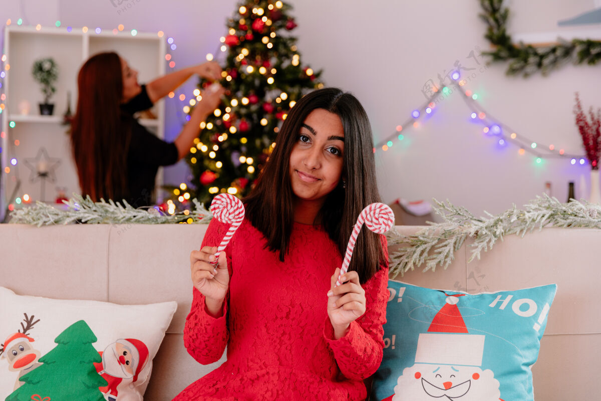 拐杖高兴的母亲装饰圣诞树和她的女儿拿着糖果手杖坐在沙发上享受圣诞节的时间在家里家圣诞树装饰