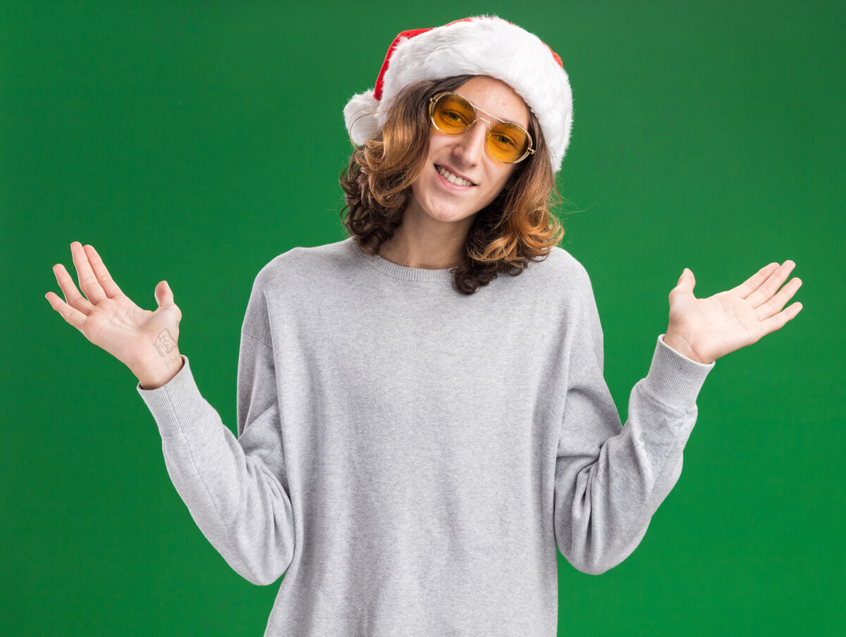 眼镜戴着圣诞老人帽 戴着黄色眼镜的年轻人面带微笑地看着摄像机 双臂伸向两侧 站在绿色背景上站立圣诞老人手臂