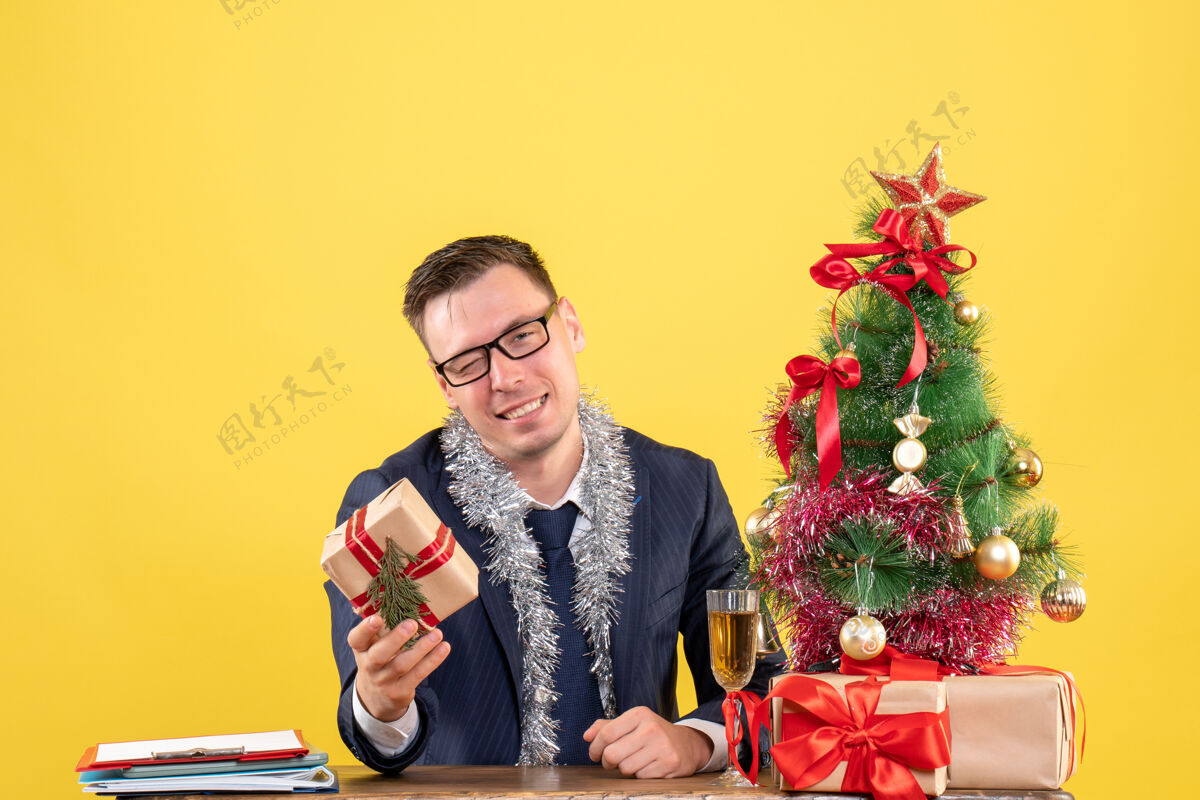 圣诞节戴着眼镜拿着礼物的快乐男人坐在圣诞树旁的桌子前 黄色的礼物成人礼物坐着