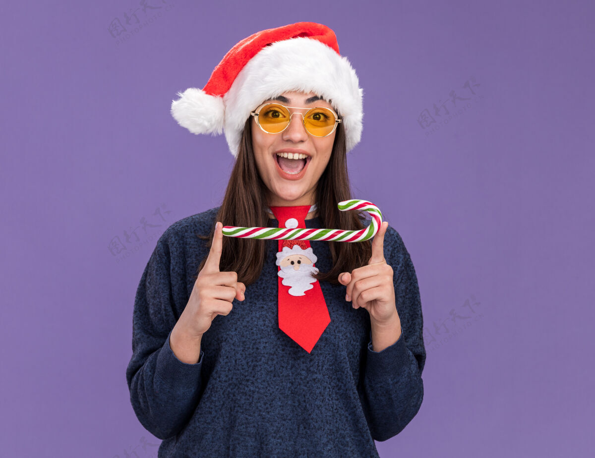 圣诞老人惊讶的年轻白种女孩戴着太阳眼镜 戴着圣诞帽 打着圣诞领带 手里拿着一根糖果棒 背景是紫色的复制空间背景快乐新的