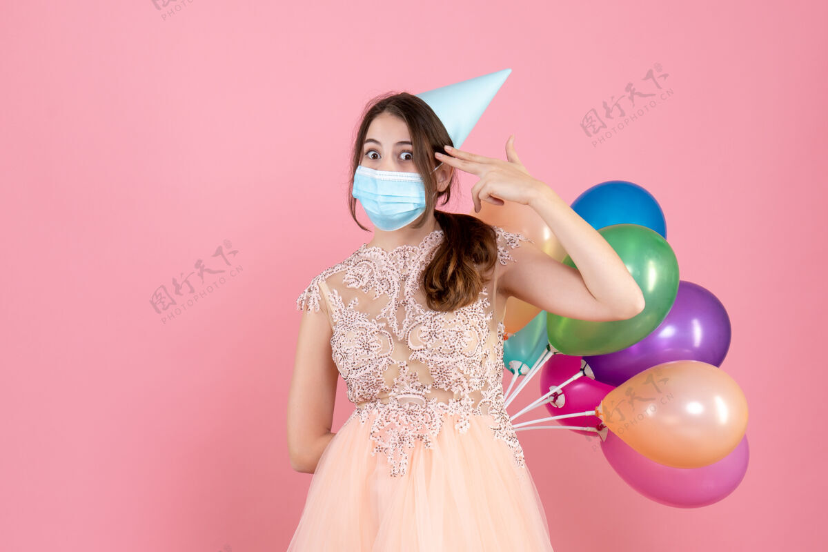 时尚戴着派对帽的大眼睛女孩背着五颜六色的气球 把手指枪放在粉红色的太阳穴附近粉色枪保持