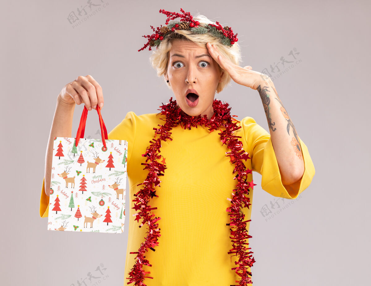 金箔惊讶的金发女郎头戴圣诞花环 脖子上戴着金属丝花环 手里拿着圣诞礼品袋 看着相机 抚摸着隔离在白色背景上的头脖子触摸包