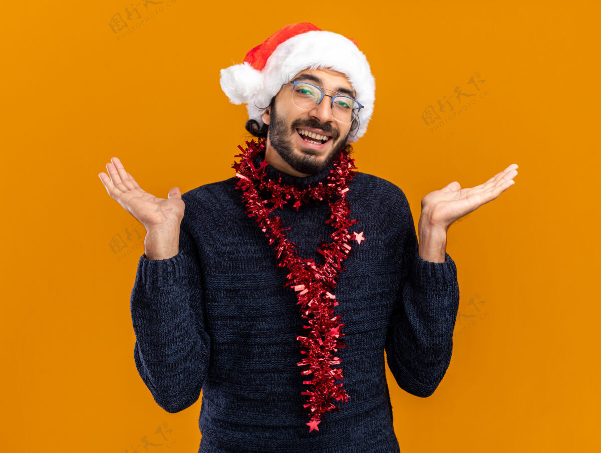年轻微笑着的年轻帅哥戴着圣诞帽 脖子上戴着花环 两手分开放在橙色的背景上帅气手蔓延