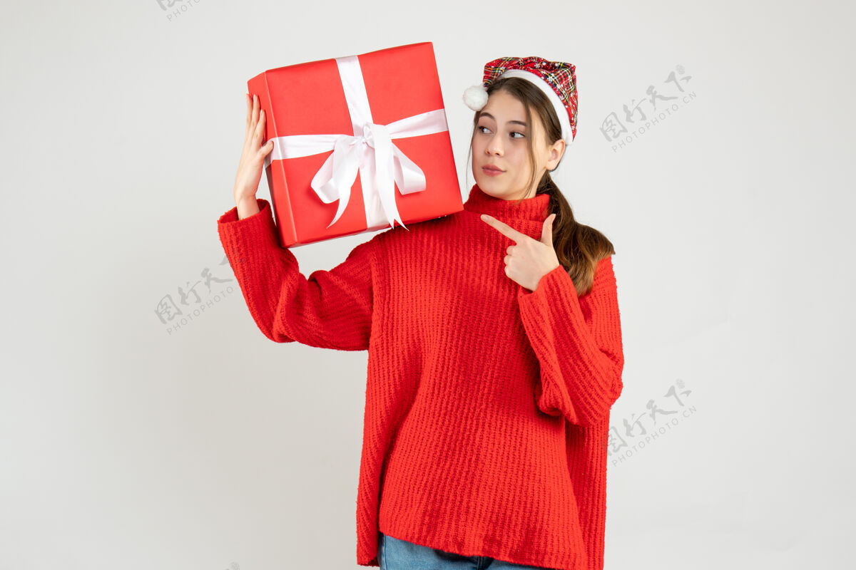 可爱戴着圣诞帽的窥探女孩拿着礼物手指着站在白色地板上的礼物手指礼物圣诞节