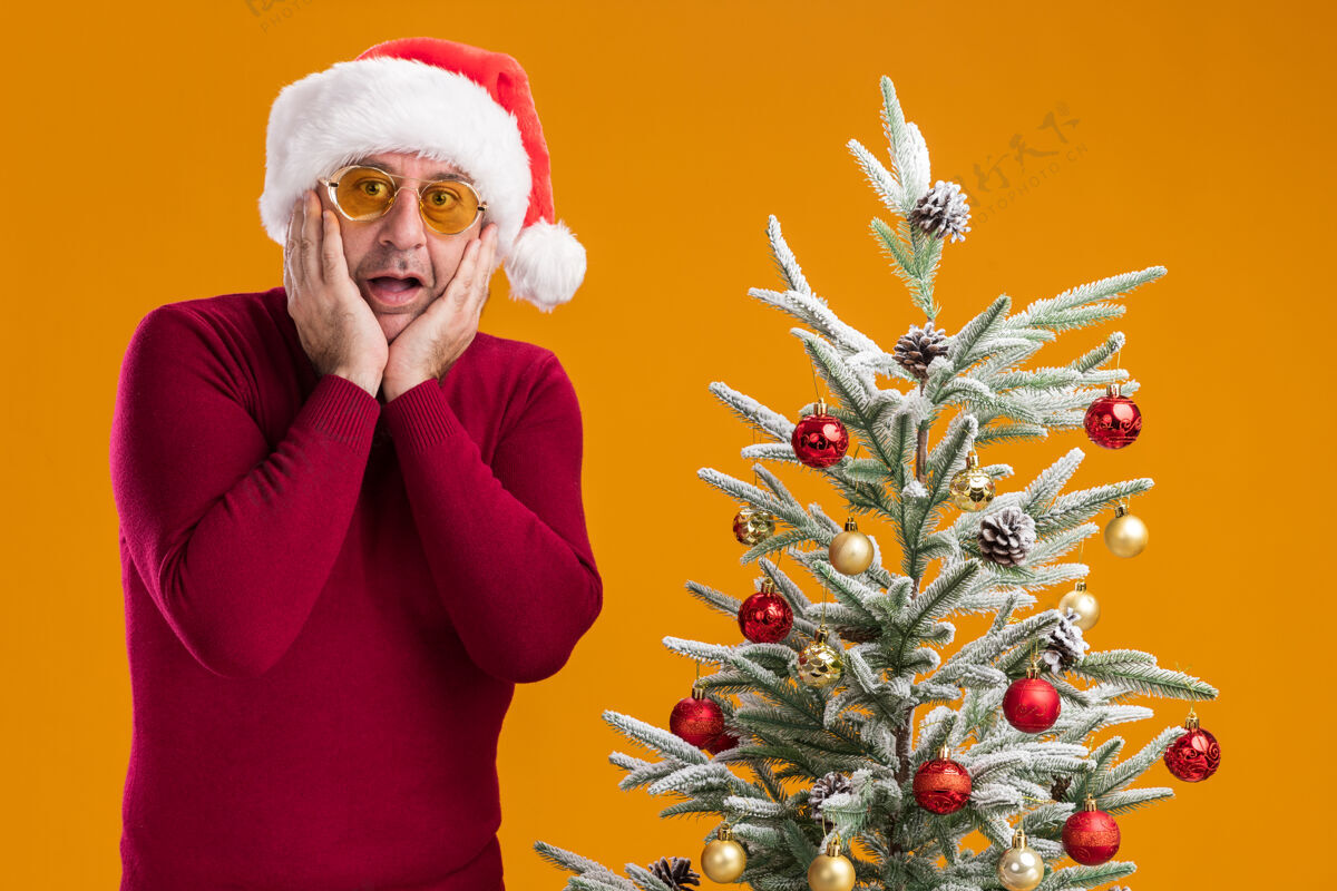 圣诞老人中年男子戴着圣诞老人帽 穿着深红色高领毛衣 戴着黄色眼镜 站在橙色背景下的圣诞树旁 惊讶地看着镜头穿着圣诞快乐眼镜