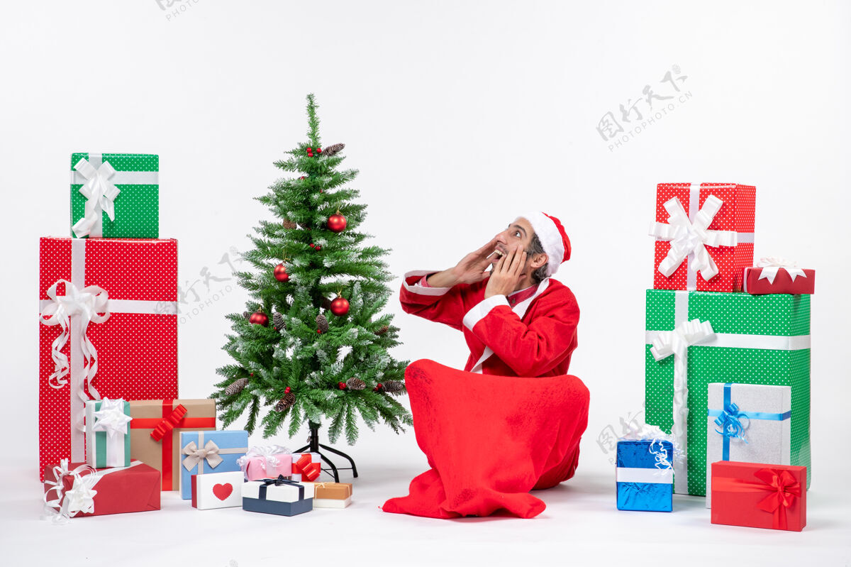 说话年轻人打扮成圣诞老人 拿着礼物和装饰好的圣诞树坐在地上 惊讶地对某人说话坐着某人地上