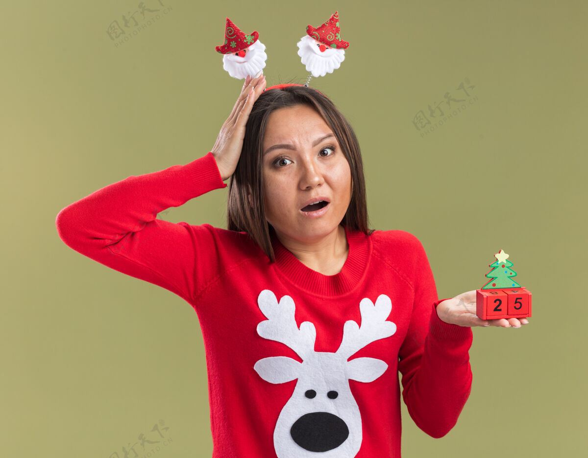 橄榄害怕的年轻亚洲女孩戴着圣诞发箍拿着圣诞玩具把手放在头上孤立在橄榄绿的墙上头圣诞节箍