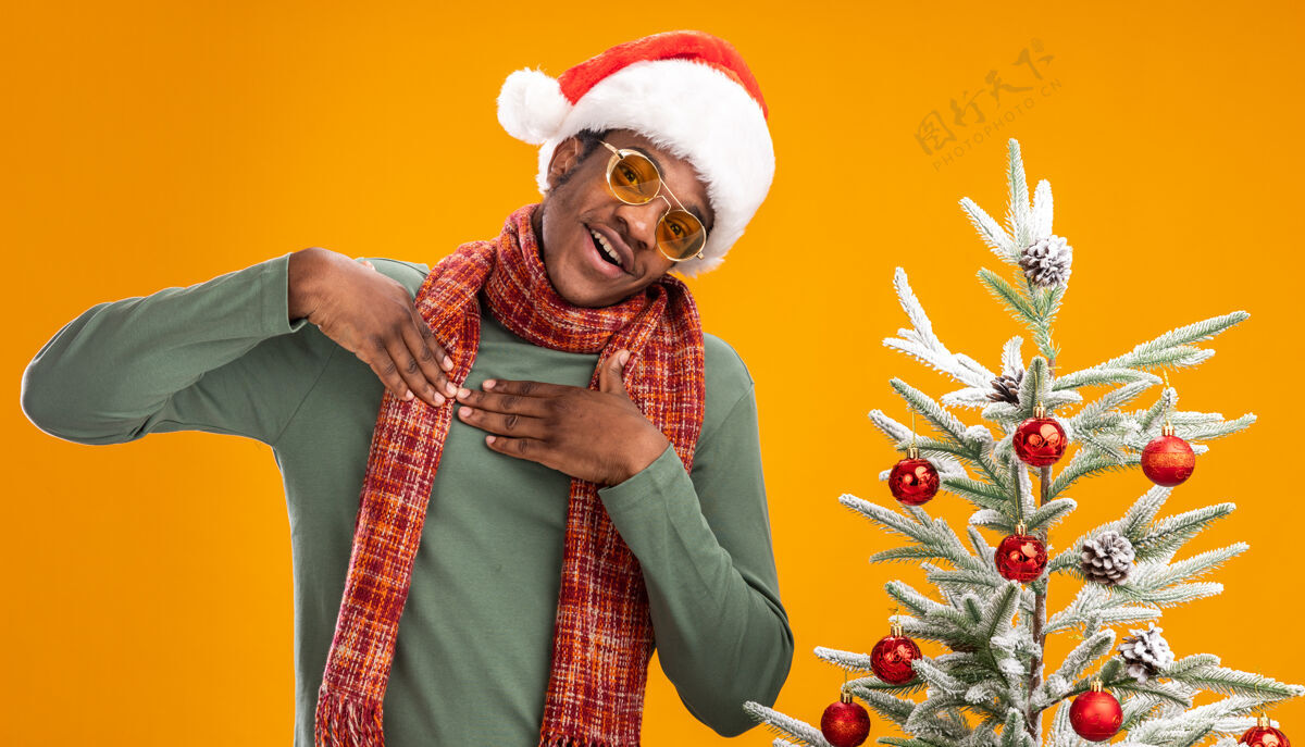站着戴着圣诞帽 围着围巾 双手放在胸前的非洲裔美国人站在橙色墙上的圣诞树旁 心情愉快 充满感激之情脖子帽子圣诞老人