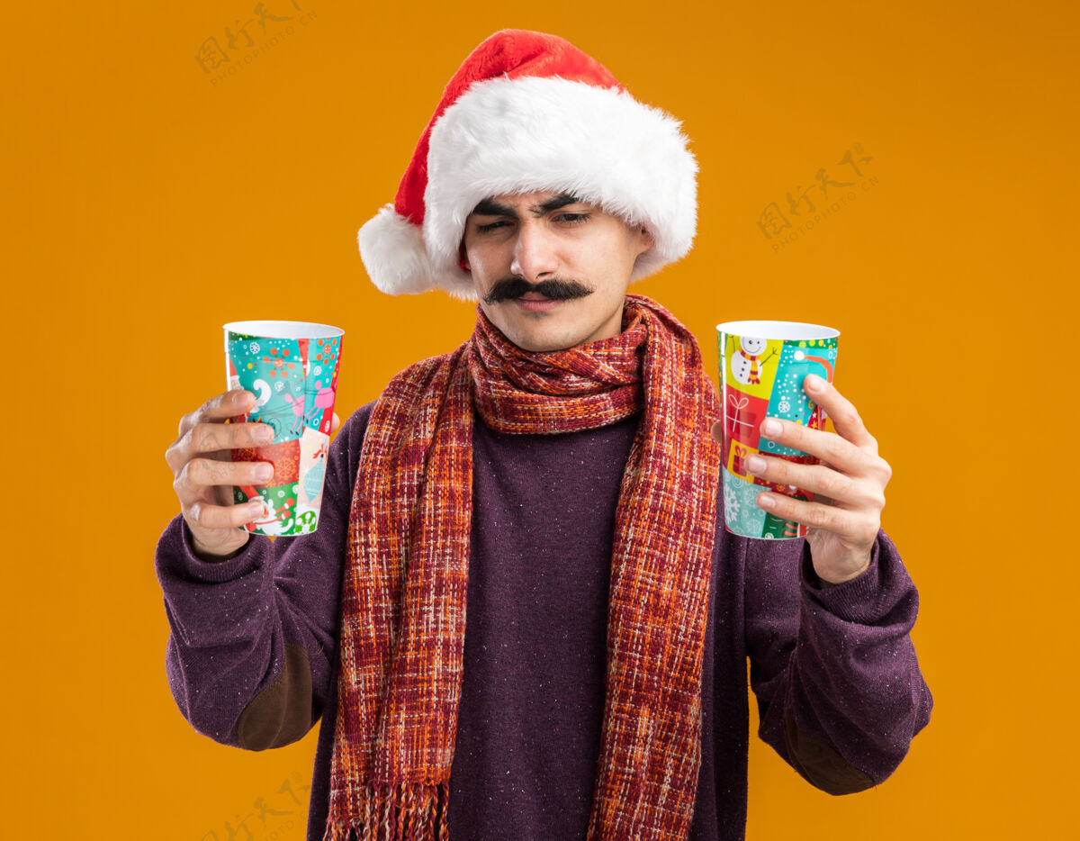 疑惑留着胡子的男人戴着圣诞老人帽 脖子上围着暖和的围巾 手里拿着五颜六色的纸杯 看起来困惑和不高兴 站在橙色的背景上有疑虑脖子橙色有