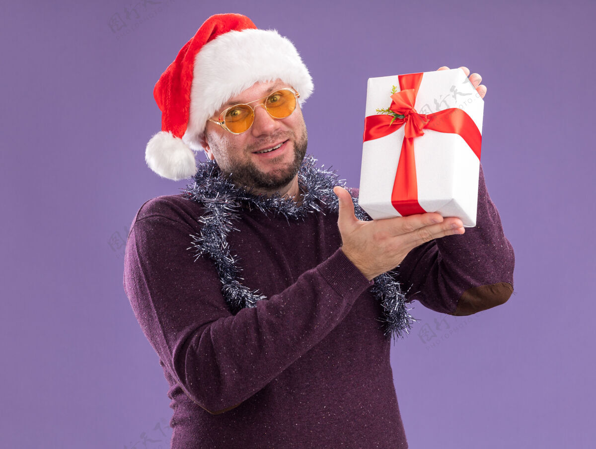 帽子微笑的中年男子戴着圣诞帽 脖子上戴着金箔花环 戴着眼镜 手里拿着礼包 隔离在紫色的墙上穿着包装圣诞节