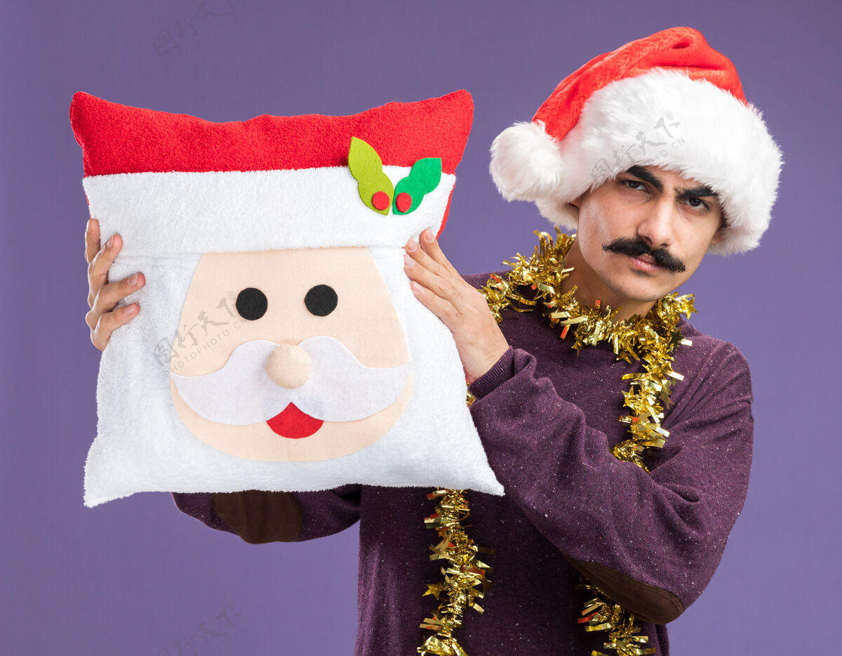 周围一个留着胡子的男人戴着圣诞帽子 脖子上戴着金属丝 手里拿着圣诞枕头 严肃地看着相机 站在紫色的背景上圣诞节脖子帽子