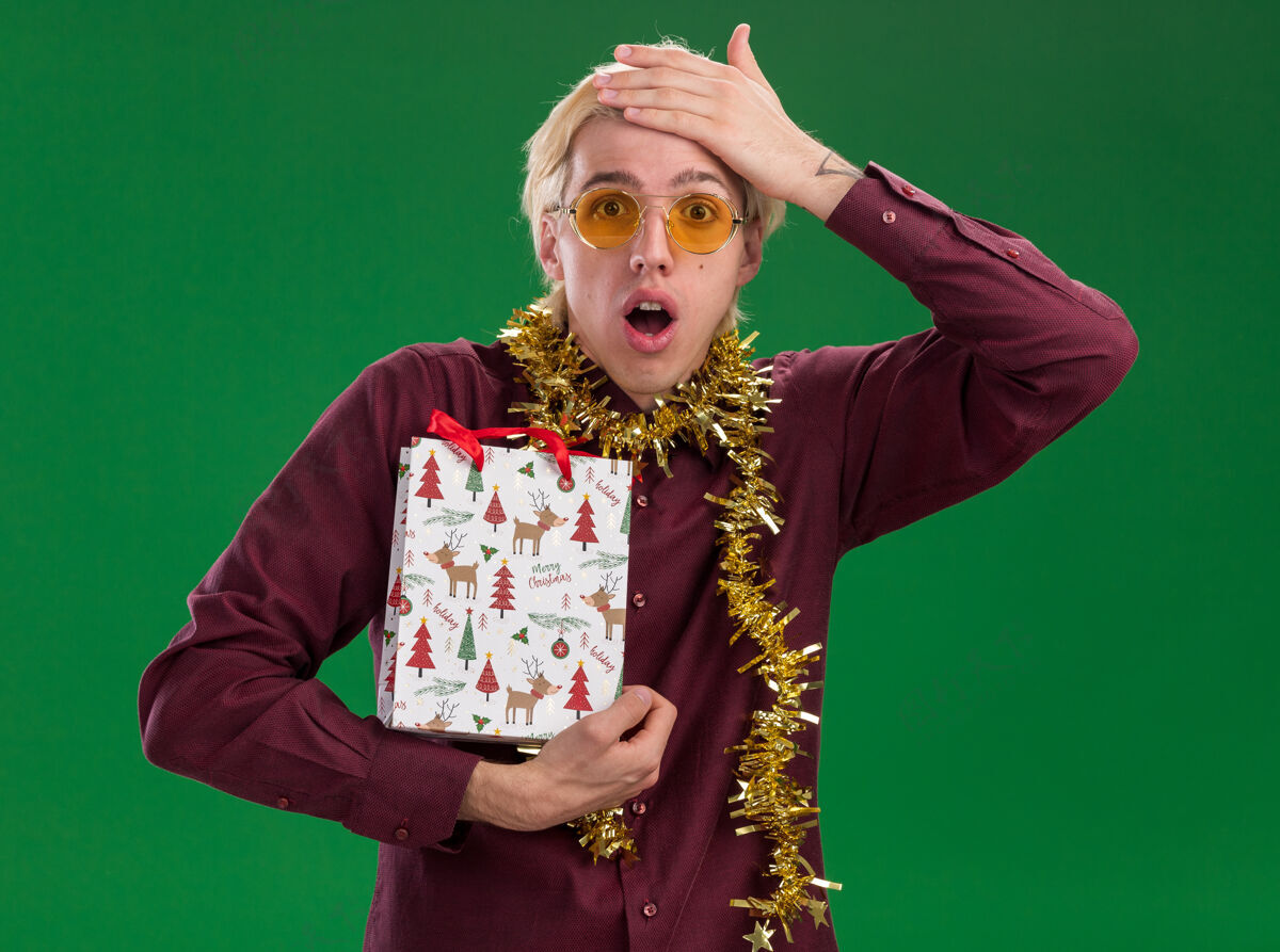 脖子遗憾的是 年轻的金发男子戴着眼镜 脖子上戴着金箔花环 手里拿着圣诞礼品袋 手放在头上 被隔离在绿色的墙上圣诞节周围遗憾