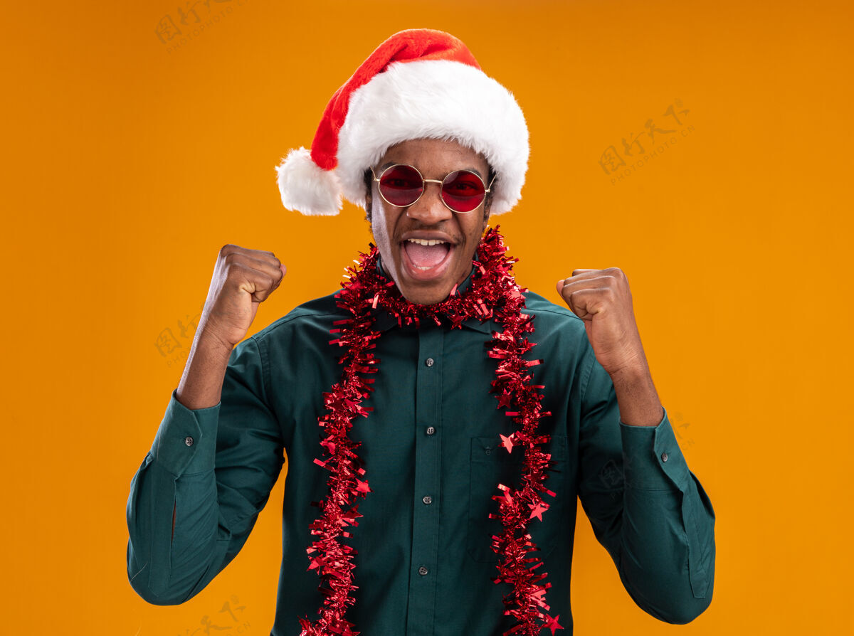 男人戴着圣诞帽 戴着花环 戴着眼镜的非裔美国人站在橙色的墙上 高兴而兴奋地握紧拳头圣诞老人花环握紧
