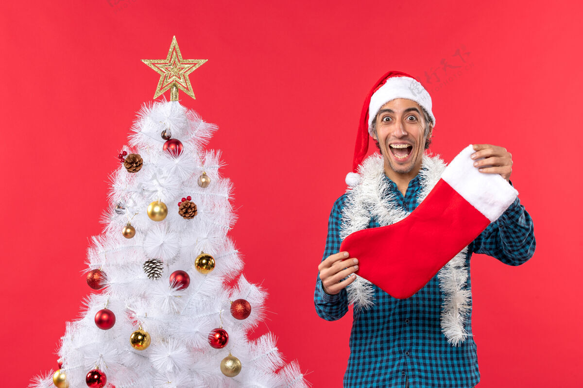 兴奋在红色圣诞树旁 一个戴着圣诞老人帽 穿着蓝色条纹衬衫 手拿圣诞袜子的激动而激动的年轻人圣诞树圣诞老人礼物