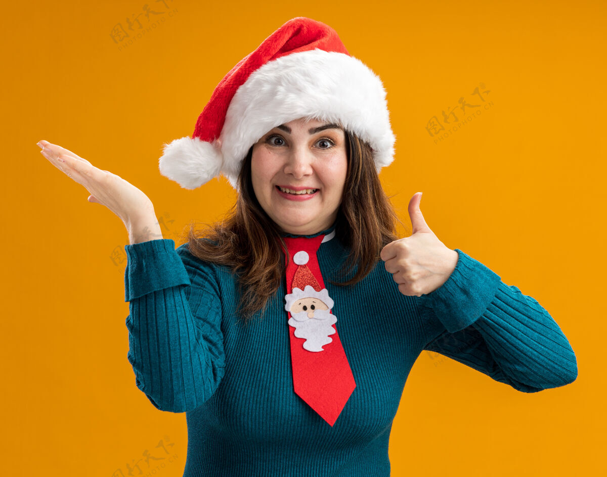 圣诞老人令人印象深刻的成年白人妇女与圣诞老人帽子和圣诞老人领带大拇指向上 并举行了手在橙色背景与复制空间隔离帽子持有拇指