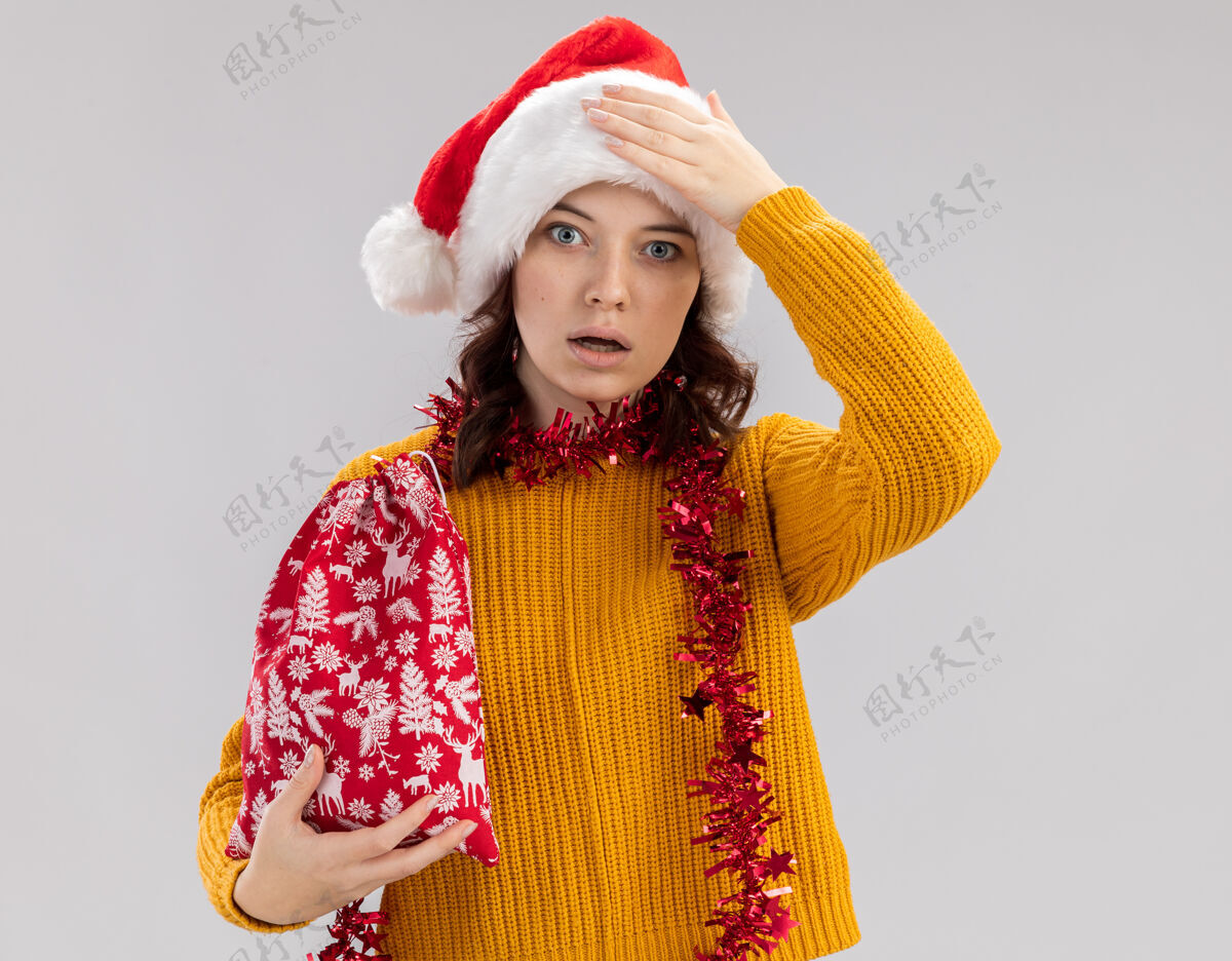 斯拉夫震惊的年轻斯拉夫女孩戴着圣诞帽 脖子上戴着花环 手放在头上 手拿着圣诞礼品袋 背景是白色的 有复制空间举行脖子花环