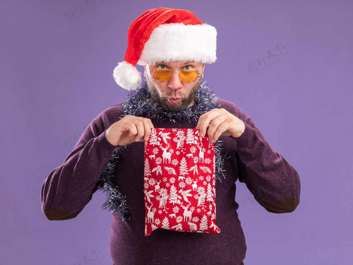 开口好奇的中年男子戴着圣诞帽 脖子上戴着金箔花环 戴着眼镜 手里拿着圣诞礼品袋 把它孤立地打开在紫色的墙上圣诞节圣诞老人好奇