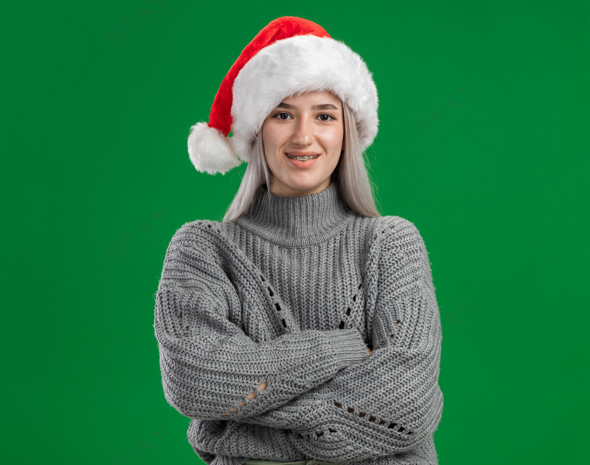 帽子年轻的金发女郎穿着冬衣 戴着圣诞帽 面带微笑 双臂交叉地站在绿色的背景上看着摄像机金发微笑圣诞老人